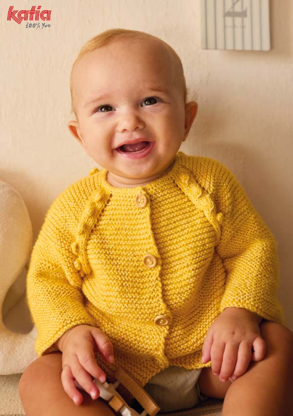 Bébé souriant portant un gilet en tricot jaune moutarde au point mousson avec boutons et détails fleuris sur les manches, assis sur un fauteuil beige.
