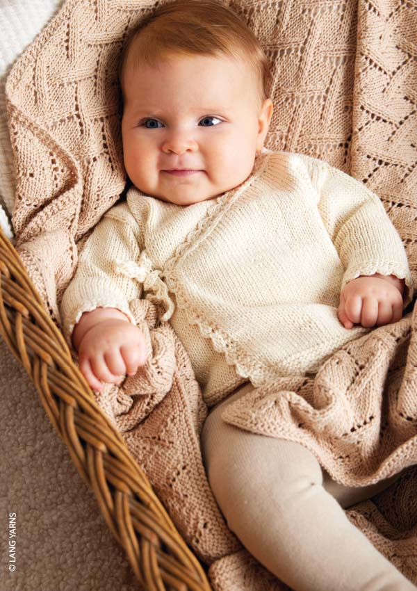 Bébé allongé dans un panier en osier, vêtu d'un cardigan en tricot ajouré écru avec finitions en dentelle, sur une couverture tricotée beige assortie.